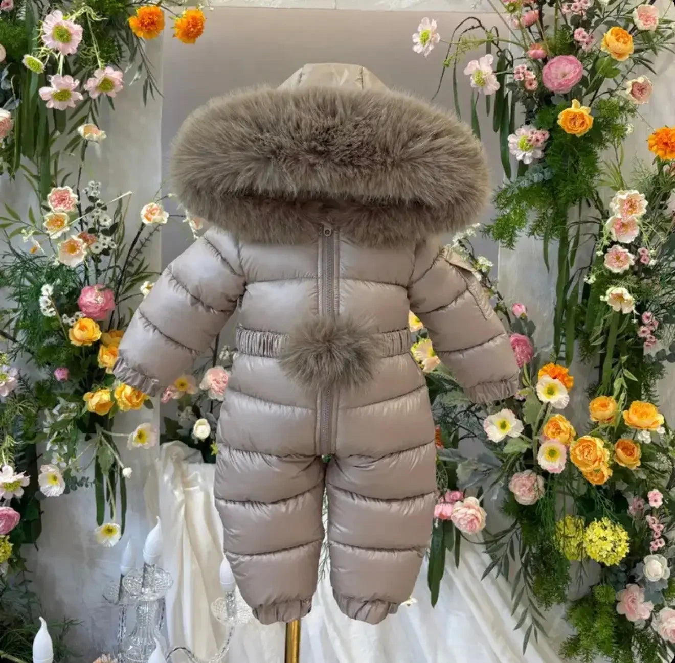 Beige padded baby snowsuit with fur trim hood and pompom, surrounded by colorful floral backdrop