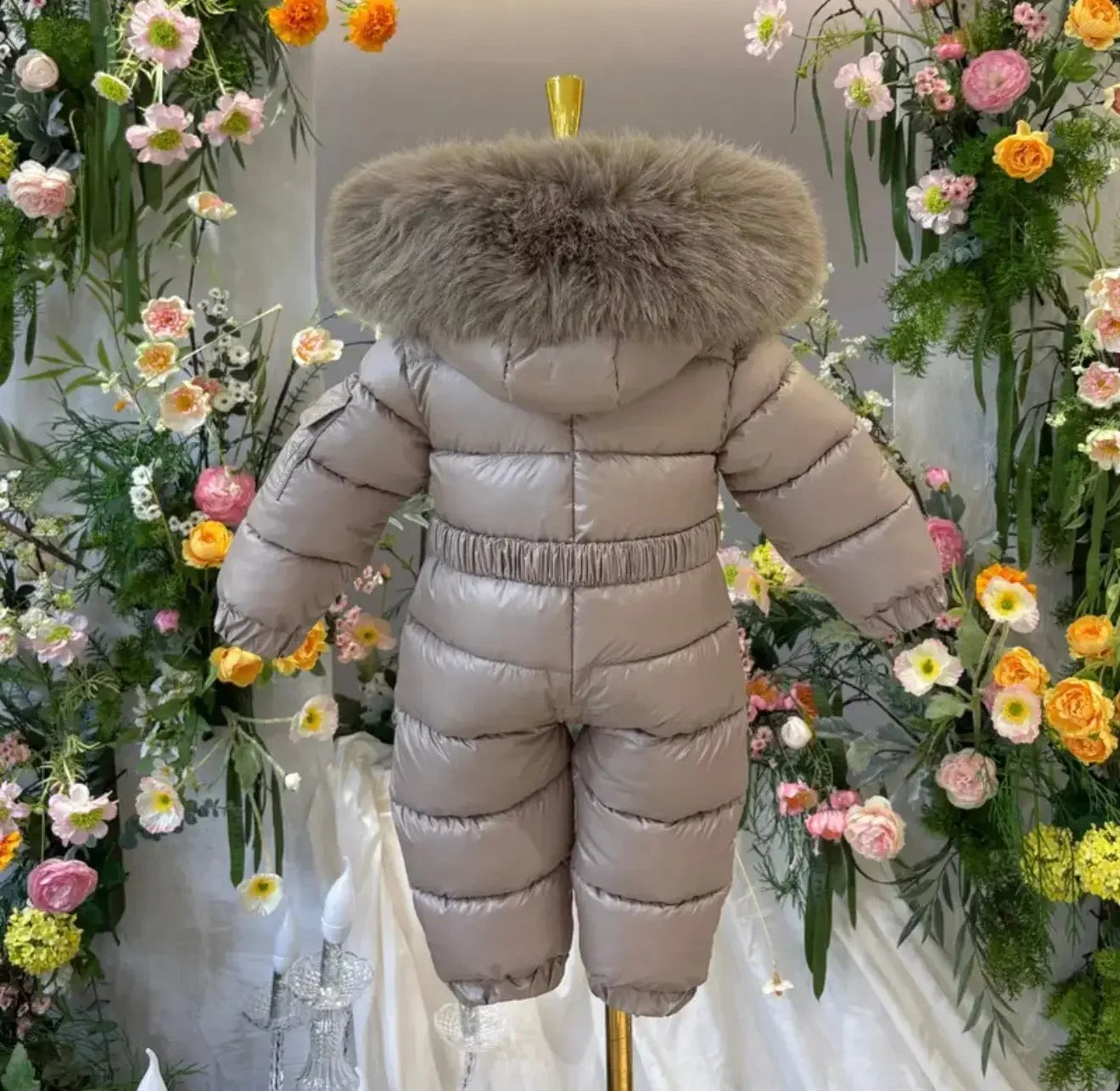 Light taupe padded baby snowsuit with large fur hood, displayed on a gold stand, surrounded by colorful flowers