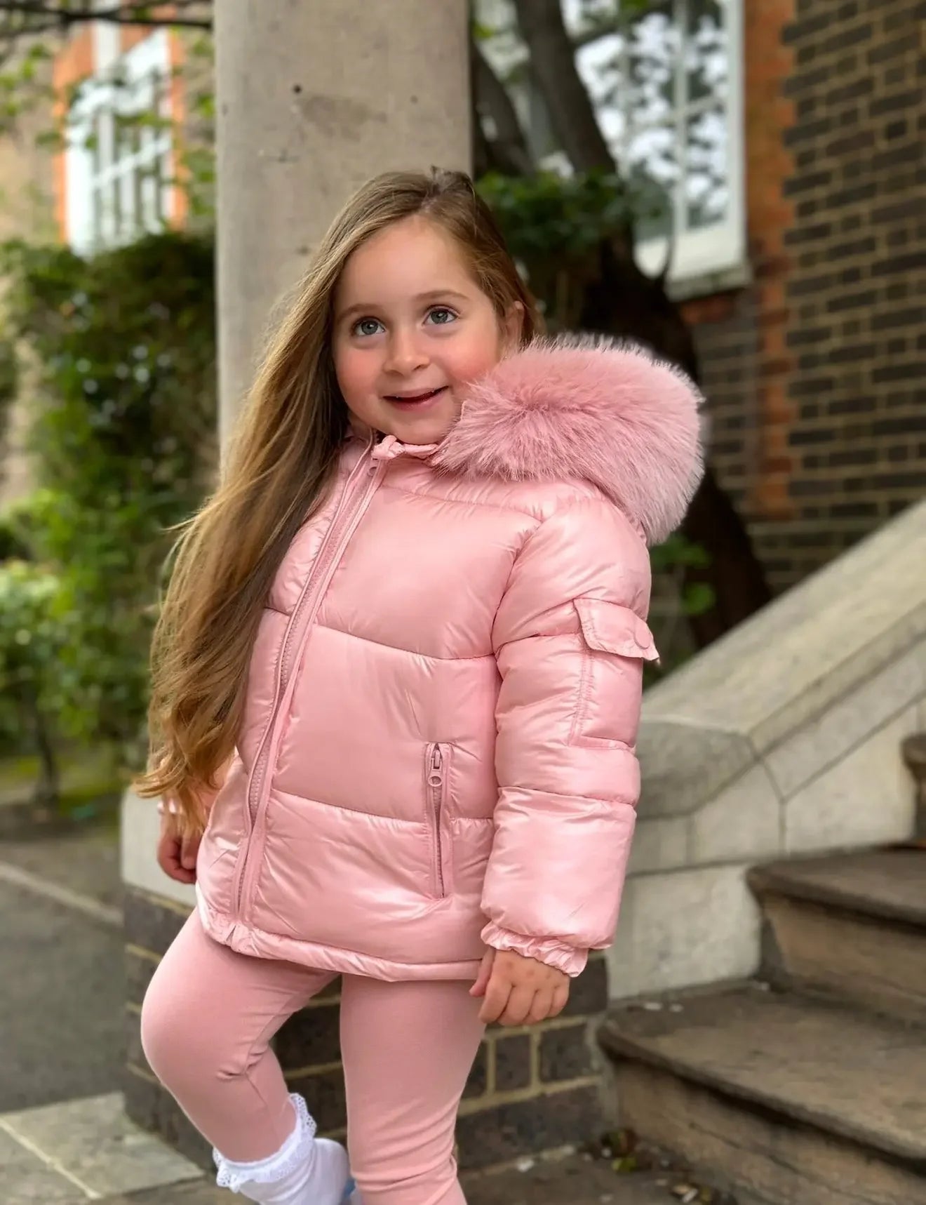 Smiling young girl wearing pink puffer jacket with fur hood and pink leggings outdoors