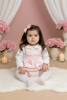 Toddler girl in white and pink outfit with bows sitting on white rug against pink backdrop with flowers and candles