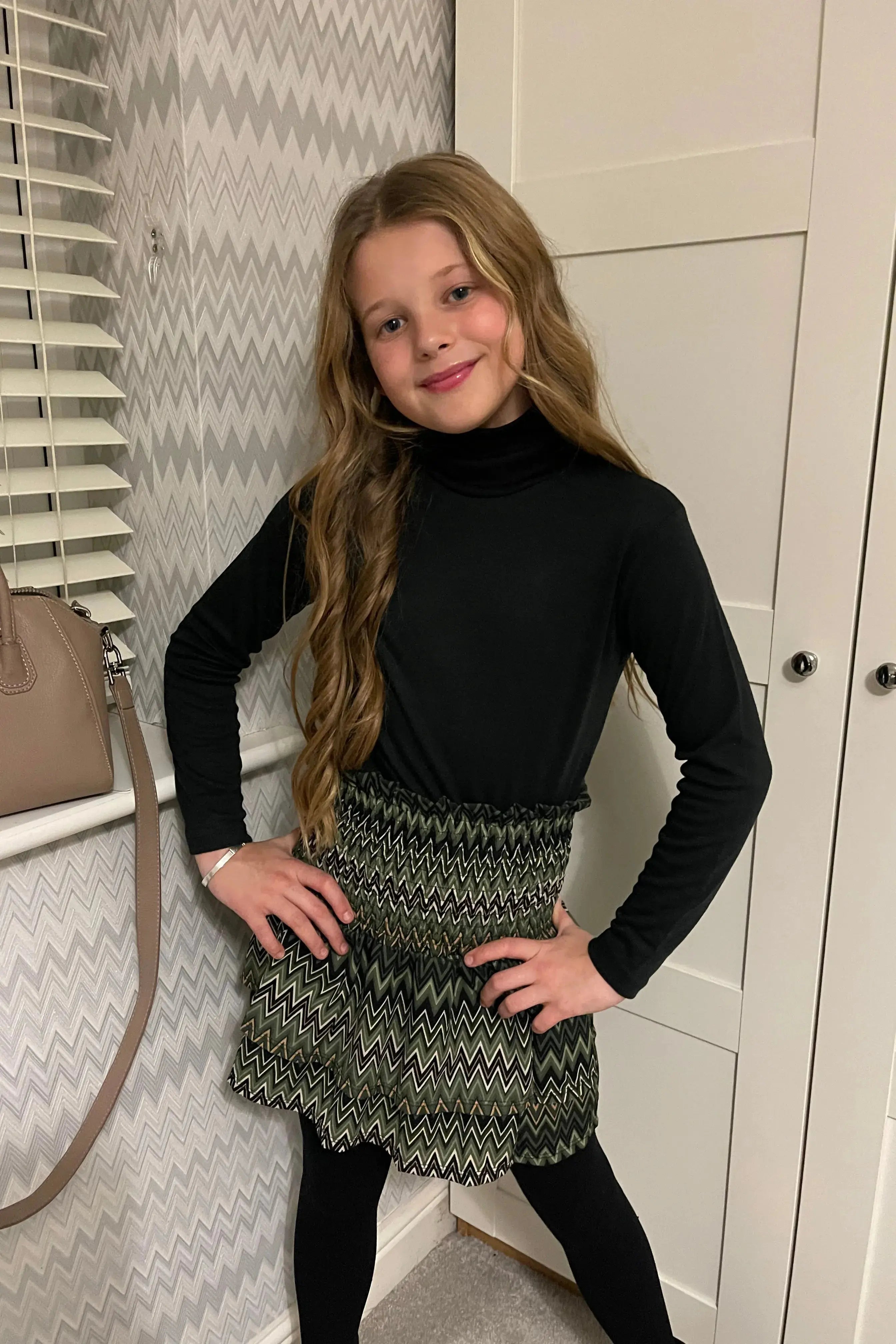 Young girl with long wavy hair wearing black turtleneck and patterned green zigzag skirt indoors
