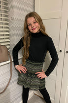 Young girl with long wavy hair wearing black turtleneck and patterned green zigzag skirt indoors