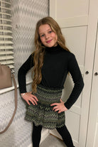 Girl in black turtleneck and green zigzag patterned skirt posing indoors with chevron wallpaper