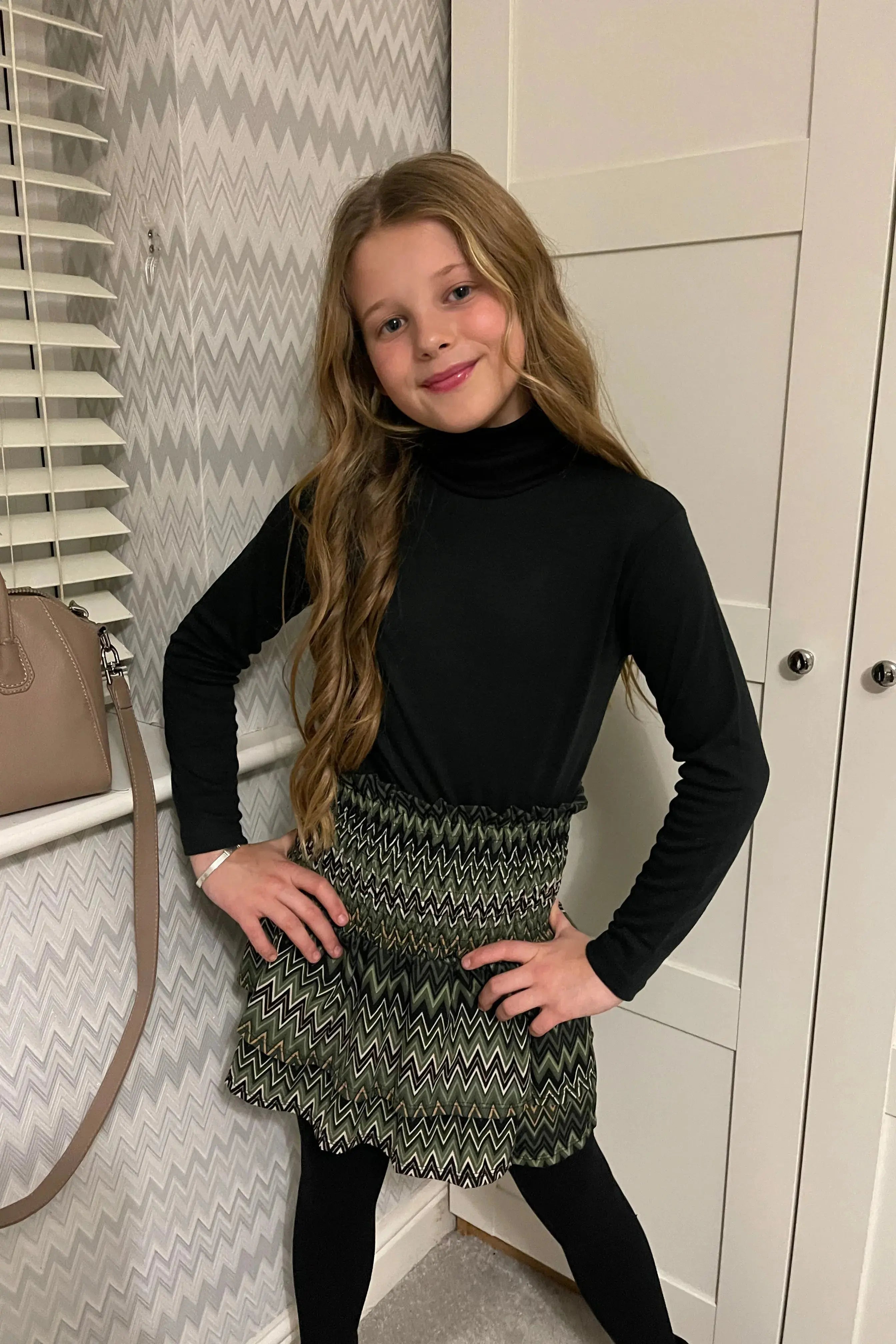 Girl in black turtleneck and green zigzag patterned skirt posing indoors with chevron wallpaper