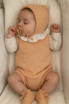 Sleeping baby in knitted peach outfit with bonnet and booties, holding pacifier, lying on soft beige bedding