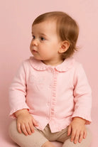 Cute baby girl in pink ruffled cardigan and beige pants sitting against pink background