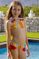 Young girl wearing a colorful yellow and red bikini with patterns, standing by a pool with greenery and stone wall backdrop