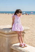 Young girl in pink dress and glittery sandals sitting on stone bench at sunny beach