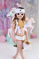 Toddler girl in beige ruffle bikini with white bows and sunglasses, holding basket, standing by pink carousel horse