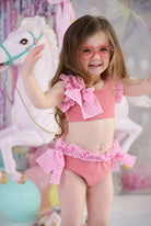 Young girl wearing pink ruffle bikini with gingham bows and pink flower-shaped sunglasses near white carousel horse