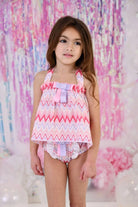Little girl wearing pink and purple zig-zag patterned bikini with bows, standing in colorful decorated room