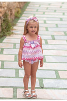 Toddler girl in pink zigzag swim top with matching headband and silver sandals on tiled path