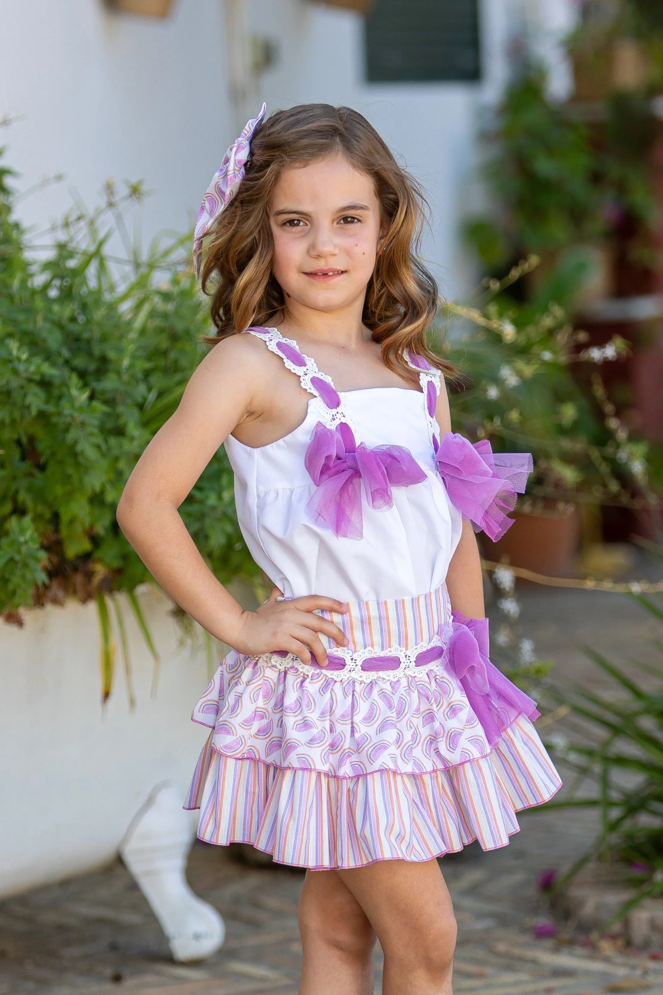 Young girl wearing a white top with purple bows and a striped tiered skirt outdoors