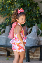 Young girl in colorful floral romper with pink bow headband posing outdoors near vintage metal containers