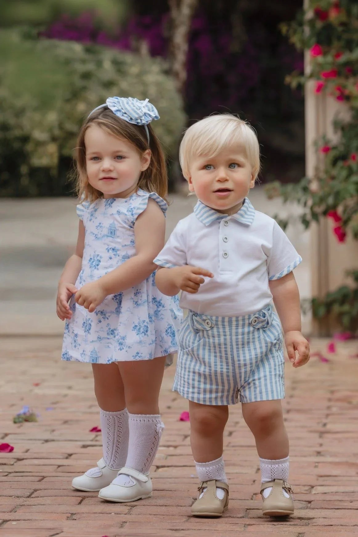 Two toddlers standing outdoors on brick pavement, girl in blue floral dress and boy in blue striped shorts