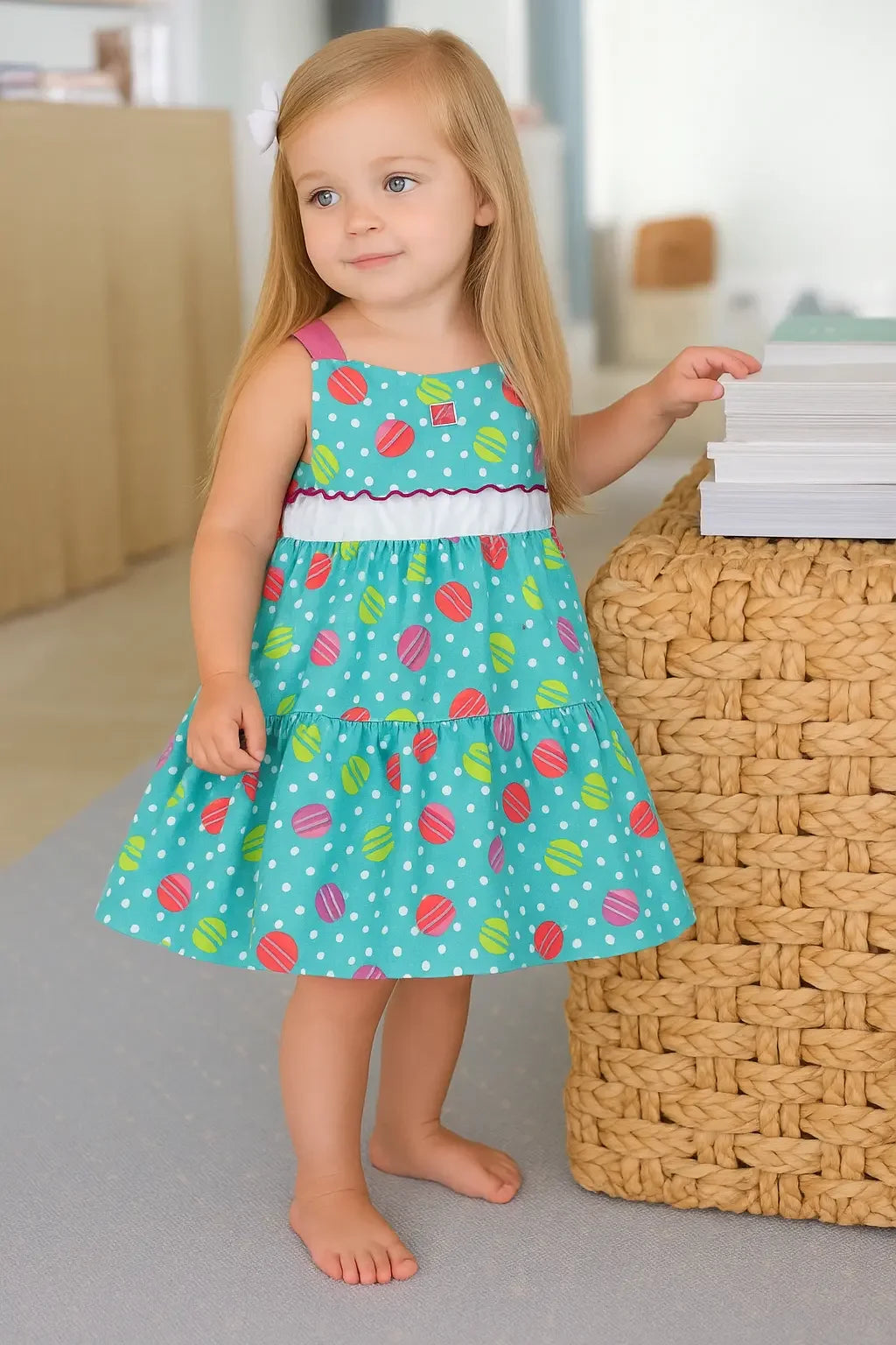Toddler girl wearing colorful polka dot sundress standing barefoot indoors beside woven basket