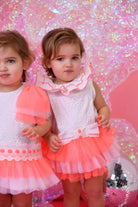 Two toddler girls wearing neon coral and white tulle dresses with ruffled collars against a pink glitter backdrop