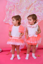 Two toddler girls in white and neon pink tulle romper dresses with white socks on a pink background