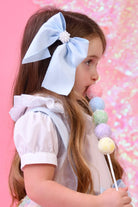 Young girl with long hair wearing a large blue bow hair clip and white ruffled blouse holding a pastel rainbow lollipop