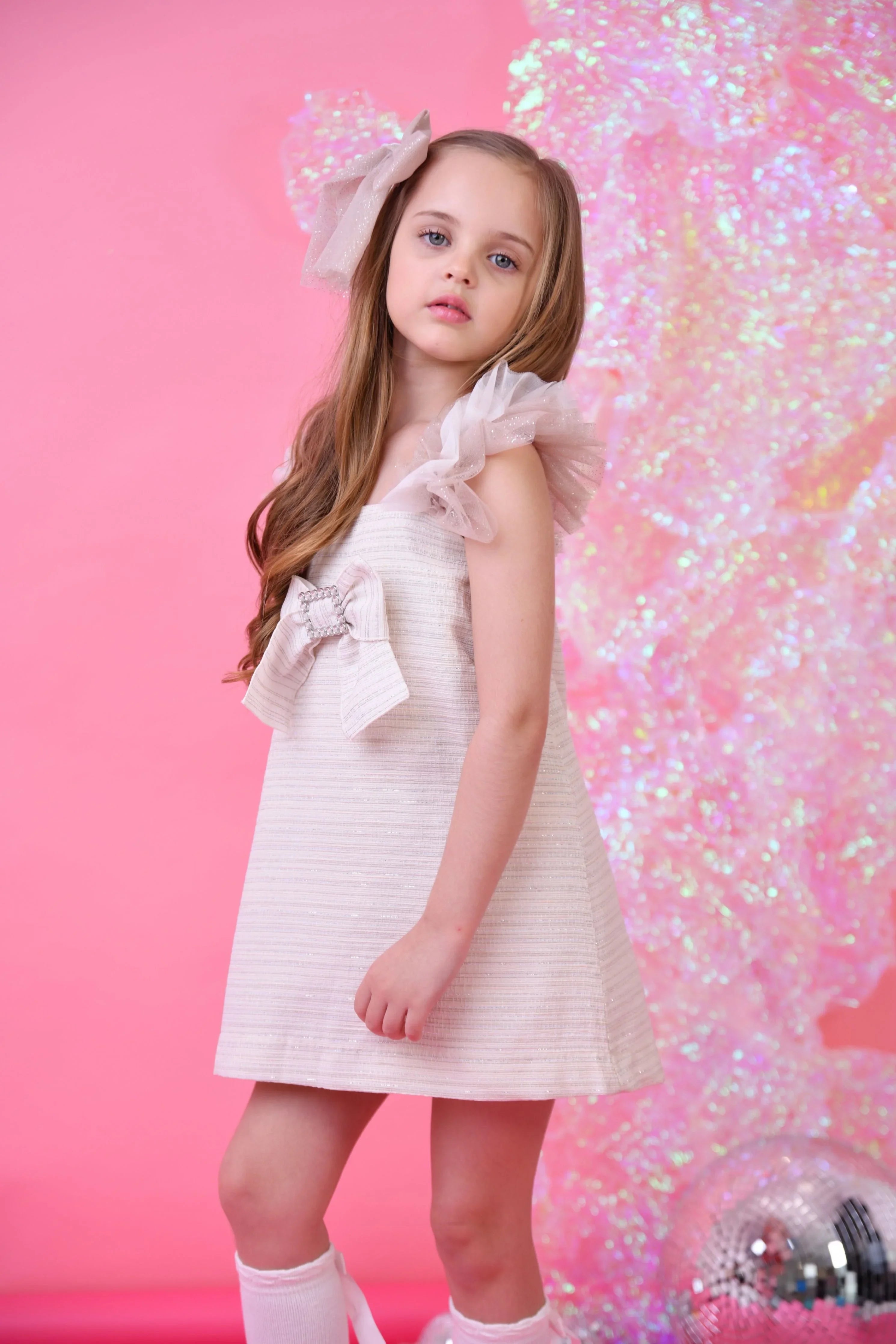 Young girl in glitter biscuit tulle dress with large bow and matching hair accessory on pink background