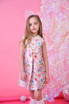 Young girl in floral sleeveless dress with pearl collar standing against pink background with sparkly decoration