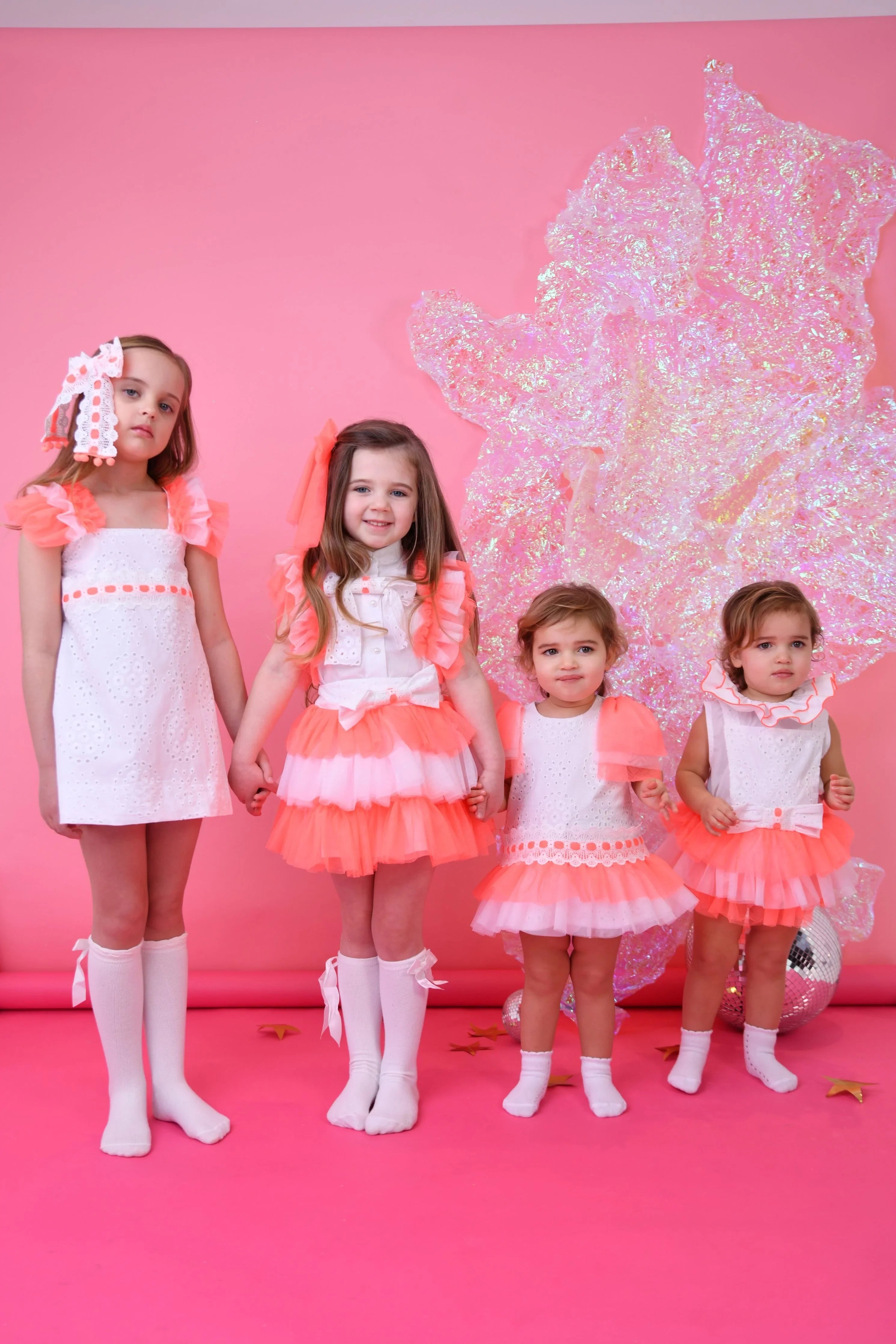 Four girls in white and neon coral tulle dresses and socks posing against a pink backdrop with iridescent decoration