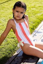 Young girl in orange and white striped swimsuit with bow detail sitting by pool on sunny grass
