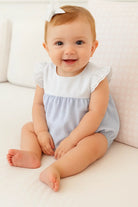 Smiling baby girl wearing a lilac ruffle romper with white bow headband sitting on white couch