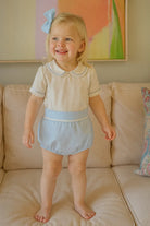 Smiling toddler girl wearing pastel blue romper with lace trim and matching hair bow on beige sofa
