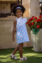 Smiling girl wearing pastel blue and pink checkered dress with white ruffle, standing outdoors near red flowers