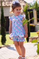 Toddler girl in pastel blue and pink checkered romper with ruffles and white bow, standing outdoors