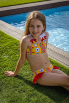 Young girl sitting by pool wearing yellow and red patterned bikini with floral shoulder details