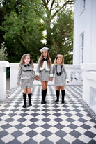 Three young girls in traditional Spanish-style grey outfits with black bows, standing on black and white checkered floor outdoors