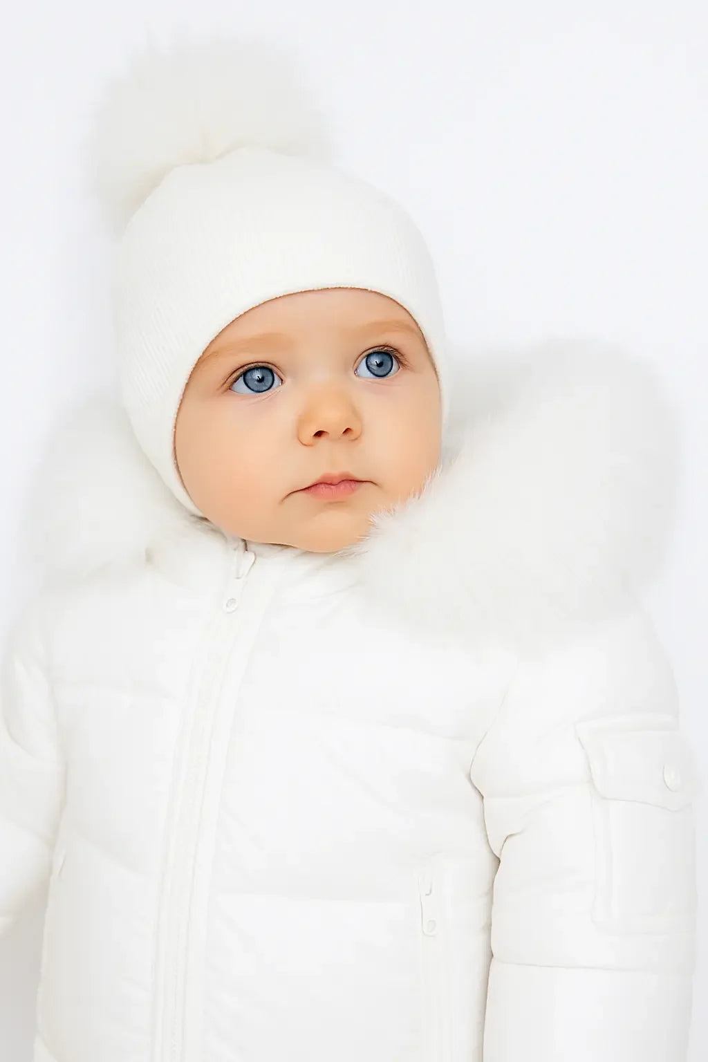 Baby with blue eyes wearing white winter jacket with fur collar and white knit hat with pompom
