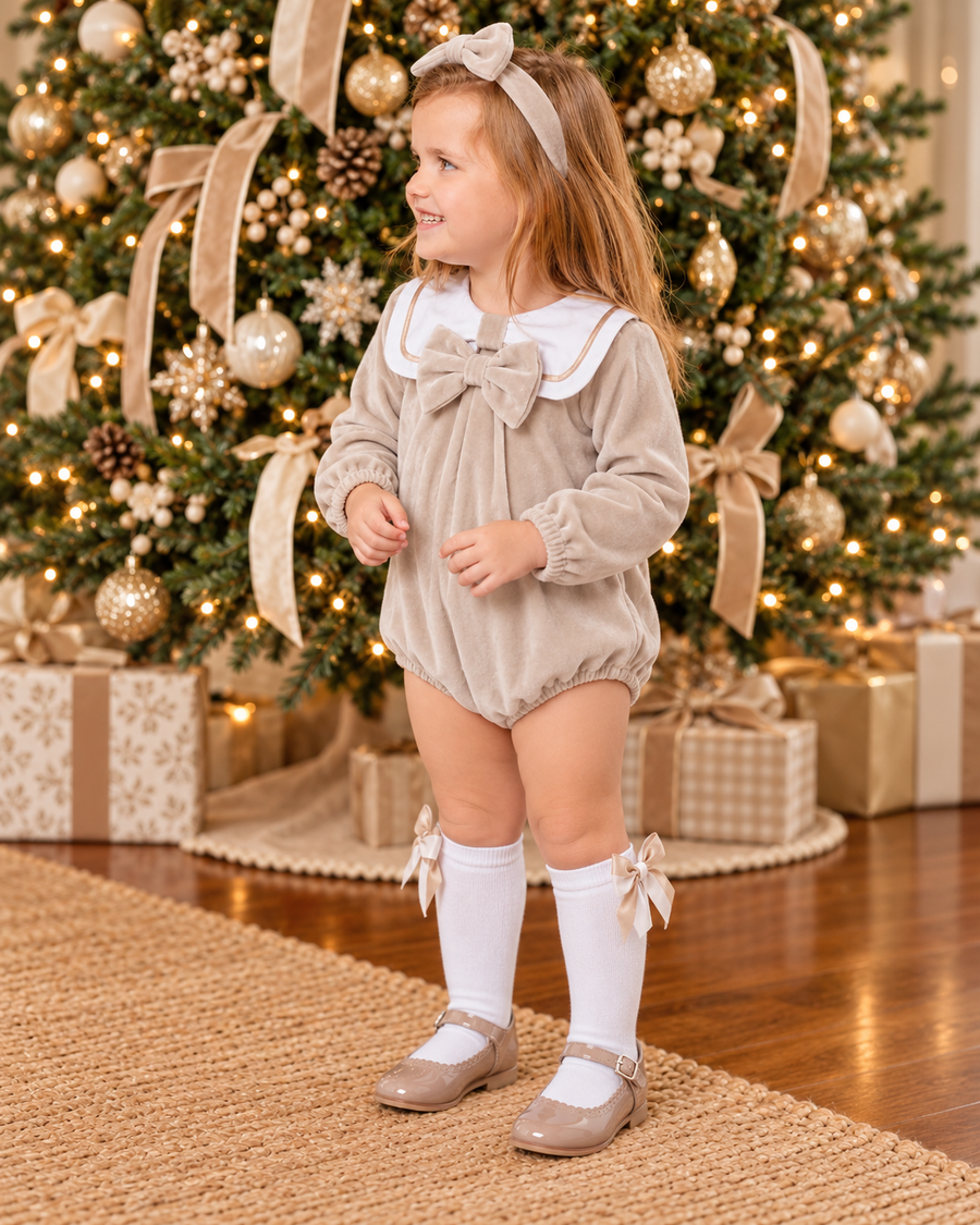 Little girl wearing a beige velour sailor collar bow romper with white knee-high socks and beige shoes in front of a beautifully decorated Christmas tree.