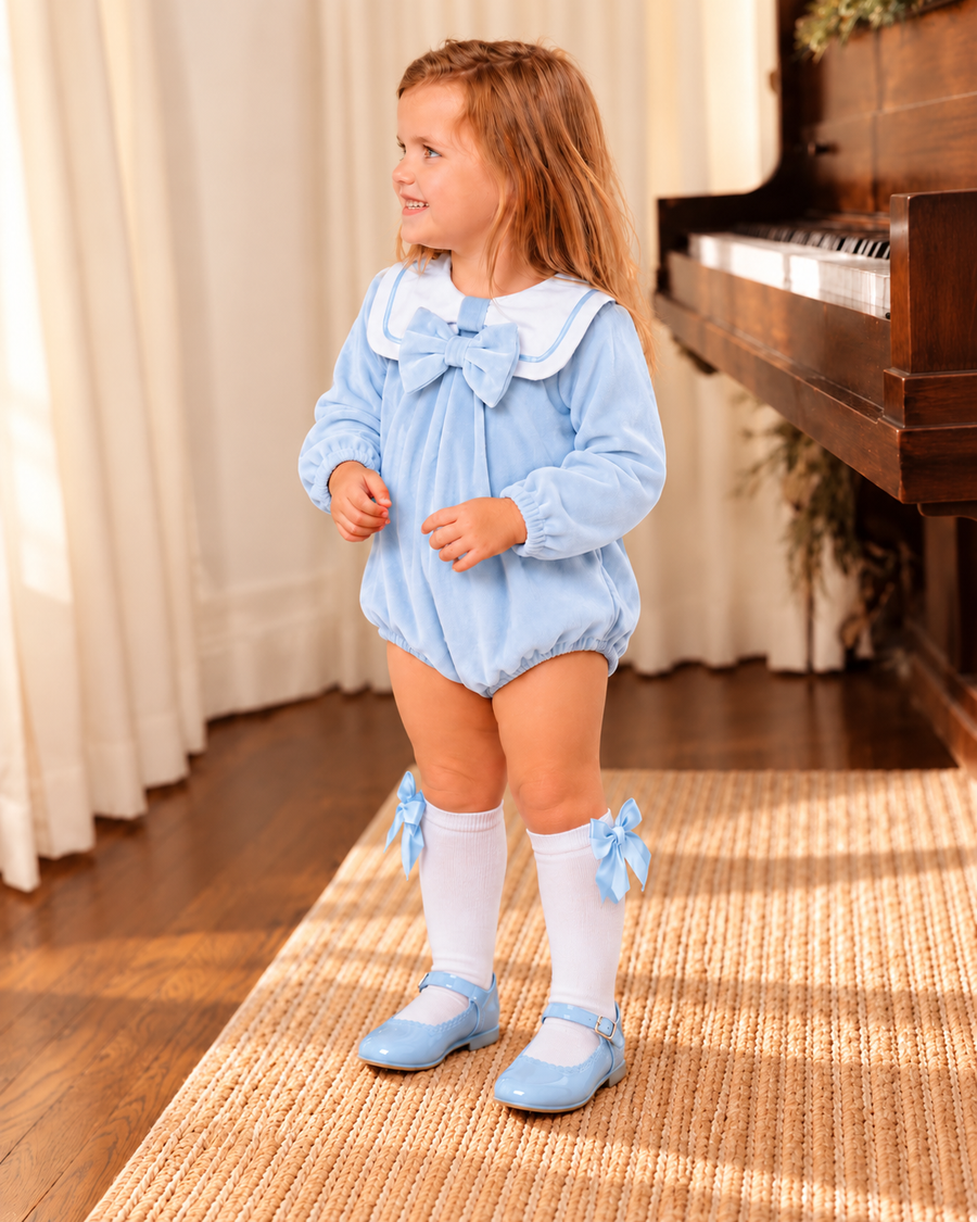 Little girl wearing a baby blue velour sailor collar bow romper with white knee-high socks and baby blue shoes beside a wooden piano.