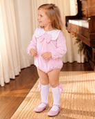 Little girl wearing a baby pink velour sailor collar bow romper with white knee-high socks and pink patent shoes beside a wooden piano.