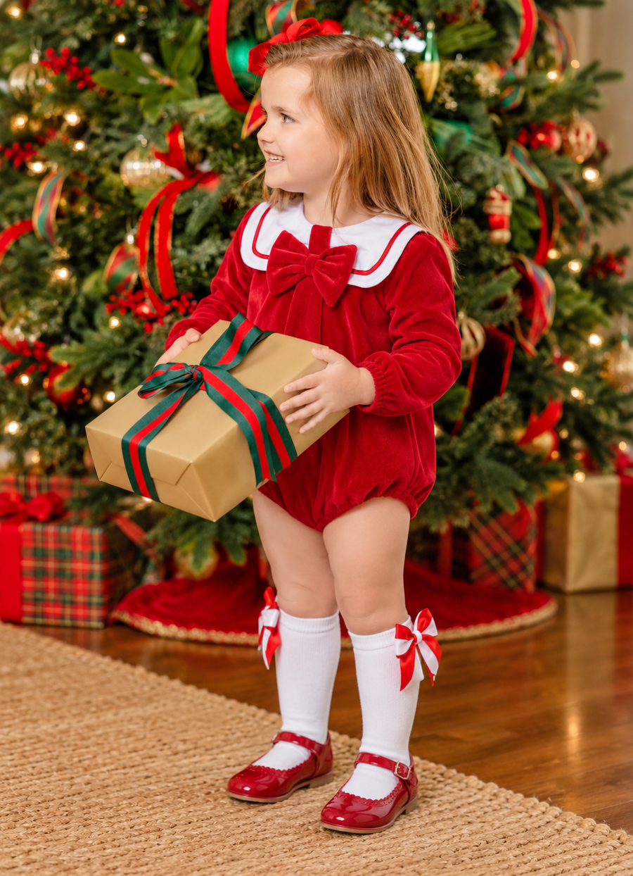 Little girl wearing a red velour sailor collar bow romper with white knee-high socks and red patent shoes beside a decorated Christmas tree.