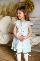 Little girl wearing a light blue sailor linen dress with cream trim, standing indoors by cozy cushions